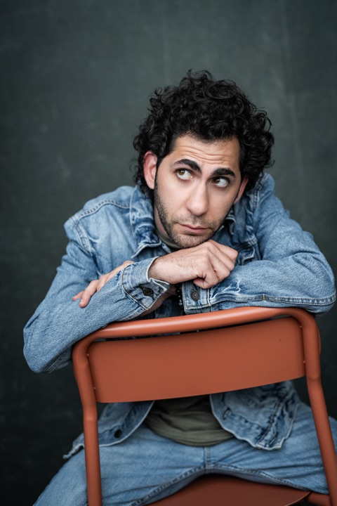 Luca Cundo in a denim jacket, glancing up over the back of an orange chair
