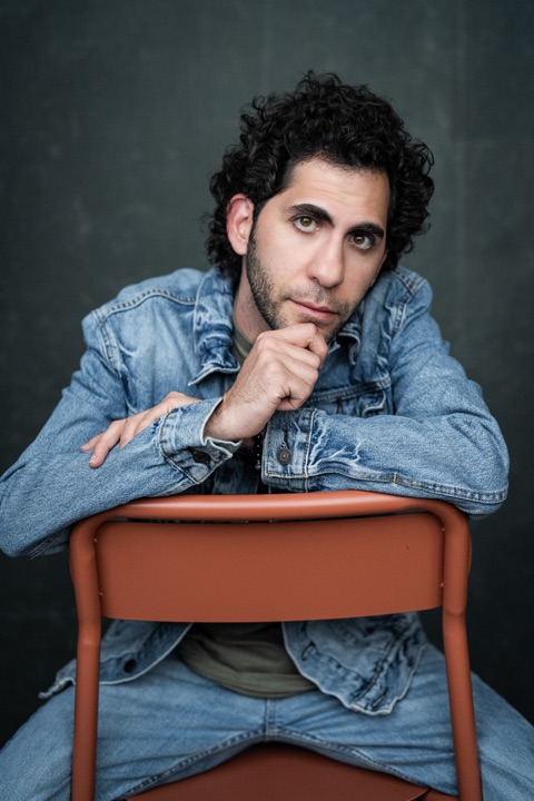 Luca Cundo in a denim jacket, hand to his chin, over the back of an orange chair