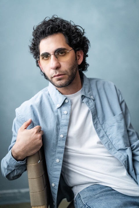 Luca Cundo in tinted glasses, seated on a chair against a blue backdrop
